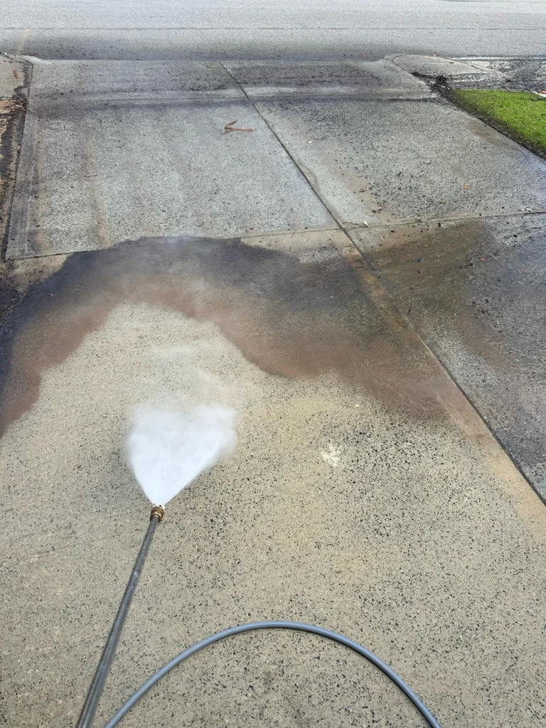 A driveway being cleaned with a pressure washer reveals striking results, its lower part sparkles whilst dark stains linger above, echoing the transformation seen in our window cleaning gallery Perth.
