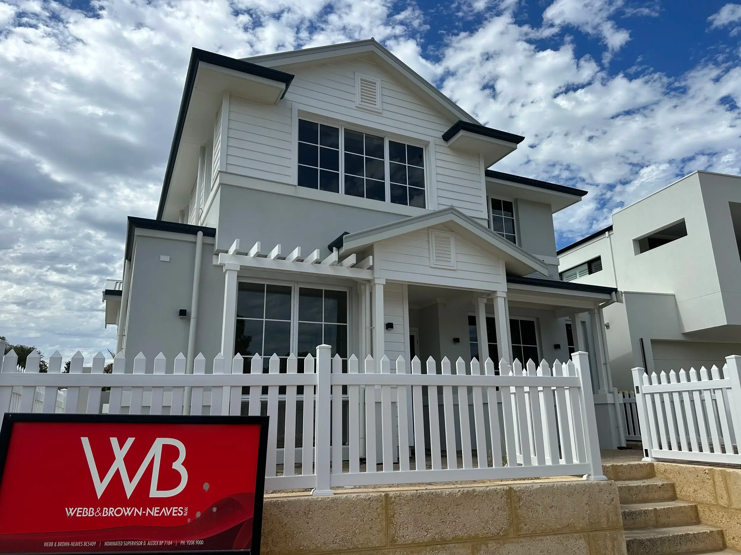 A modern two-storey white house with large windows, a white picket fence, and a red estate agent sign in front reading "WB WEBB & BROWN-NEAVES." Featured in a window cleaning gallery Perth, the partly cloudy sky frames this stunning home.