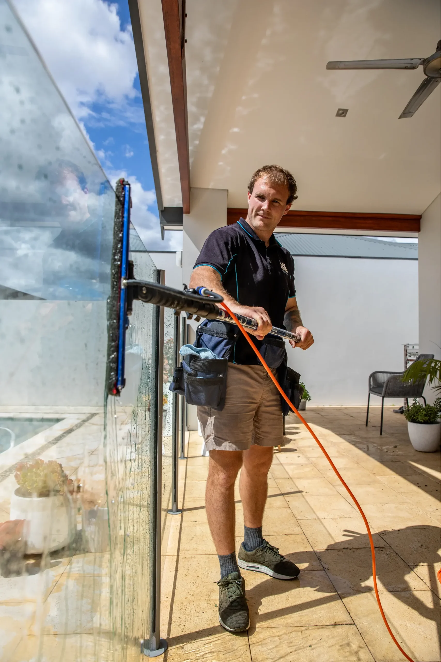 A man in shorts and a polo shirt uses a long-handled tool to clean an outdoor glass balustrade on a sunny patio, with plants and chairs in the background.