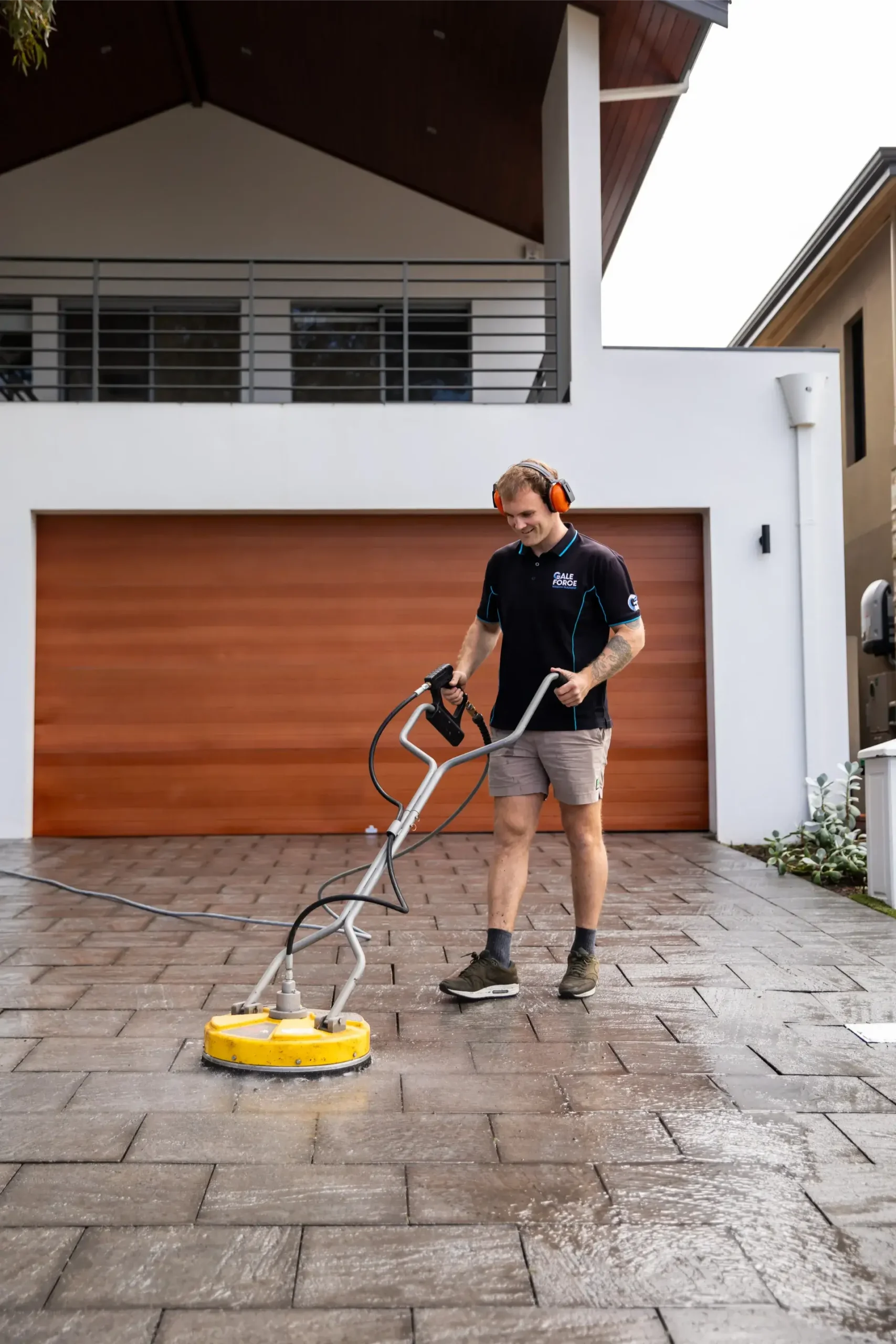A person wearing ear defenders uses a yellow surface cleaner to wash a paved drive in front of a modern house with a wooden garage door.