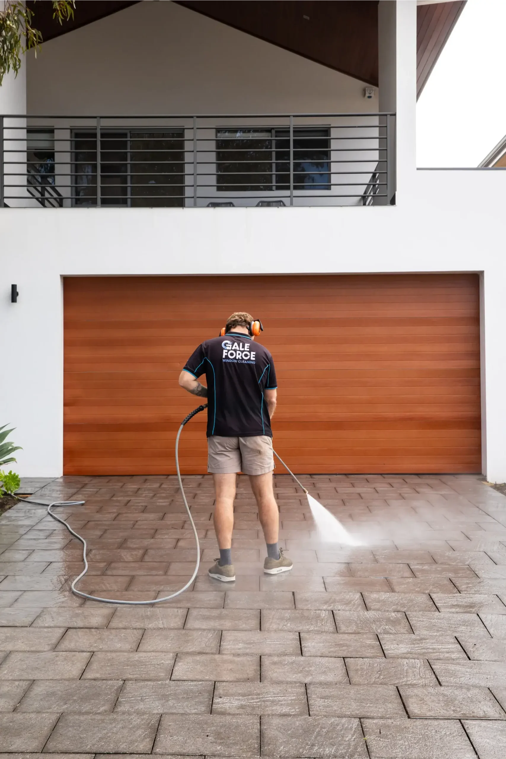 A person wearing ear protection uses a pressure washer to clean a paved drive in front of a closed wooden garage door and a modern white house.