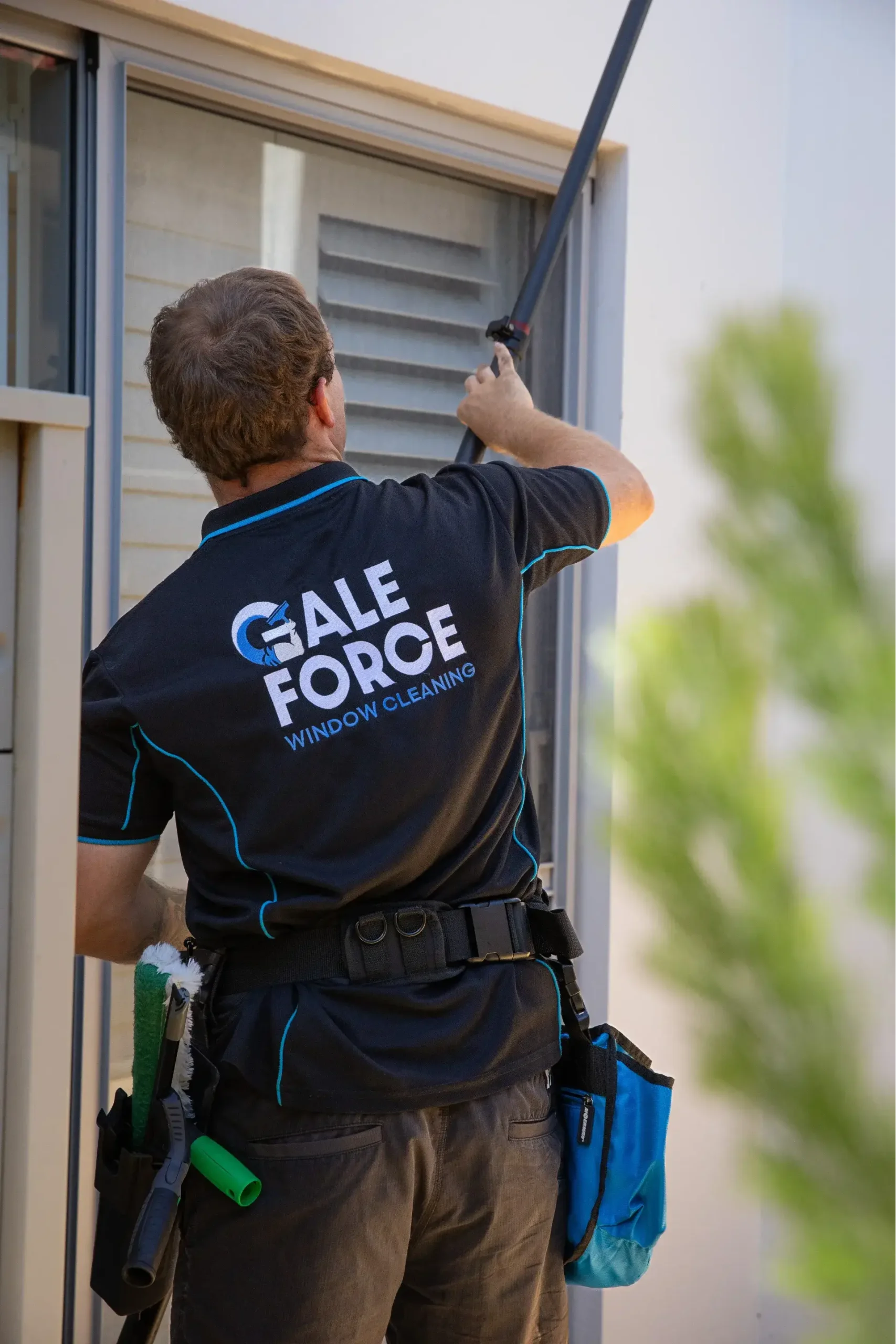 A window cleaner wearing a “Gale Force Window Cleaning” shirt uses a long pole to clean a window. Cleaning tools and pouches hang from their belt. The person is working outside a building.