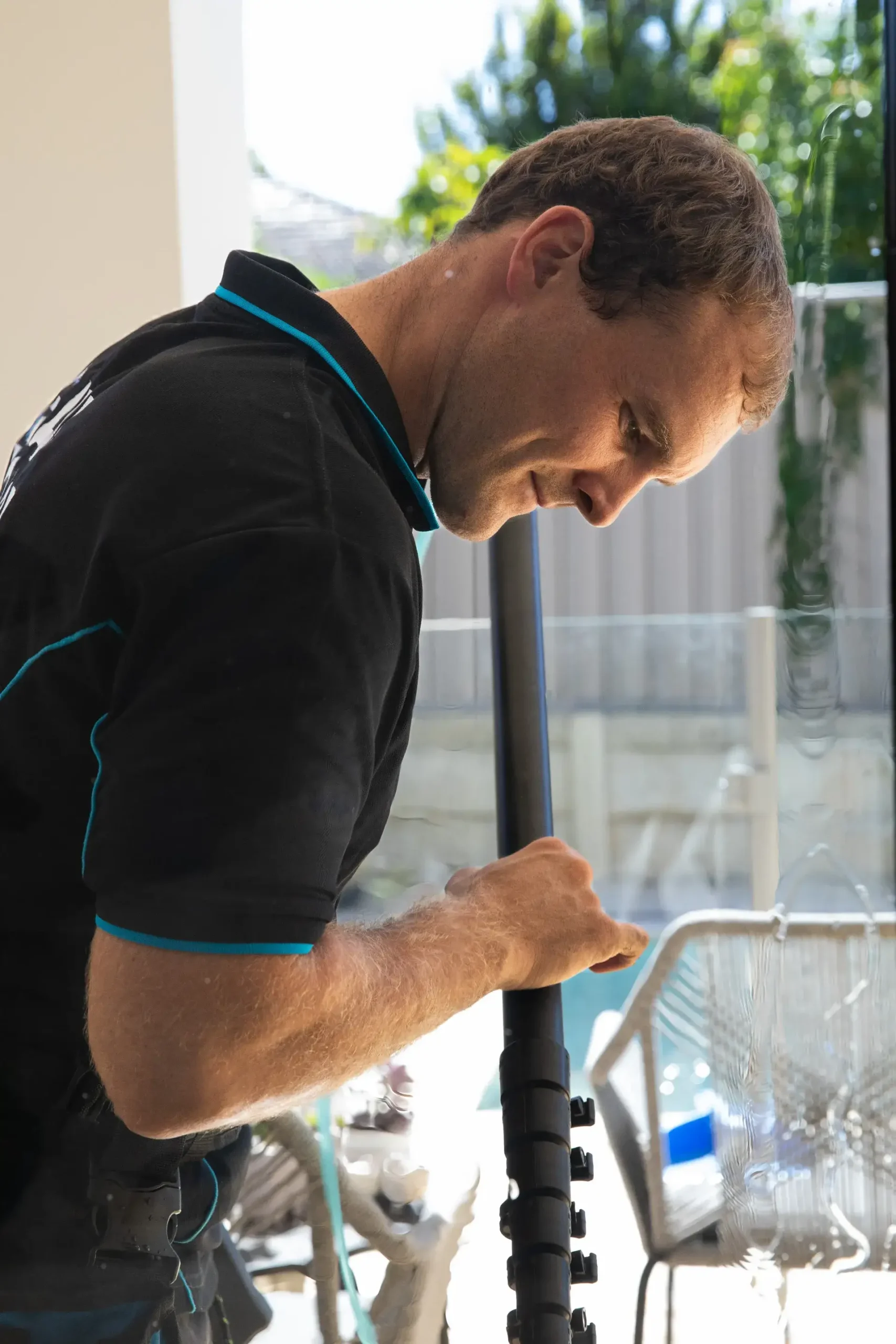 A man in a black shirt with blue trim uses a cleaning tool to wash a glass window, with outdoor furniture and greenery visible in the background.