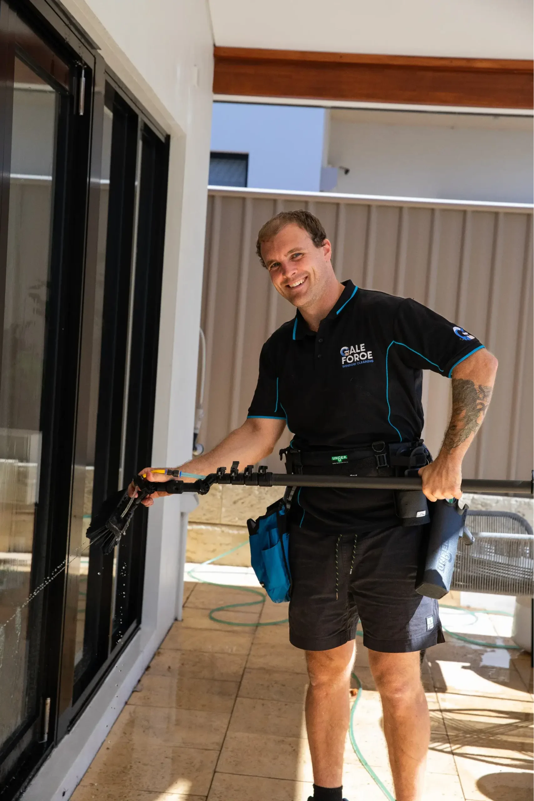 A smiling man in a black uniform is cleaning a glass door with a water-fed pole. He stands on a patio with tiled flooring, holding cleaning equipment, and wears a tool belt round his waist.