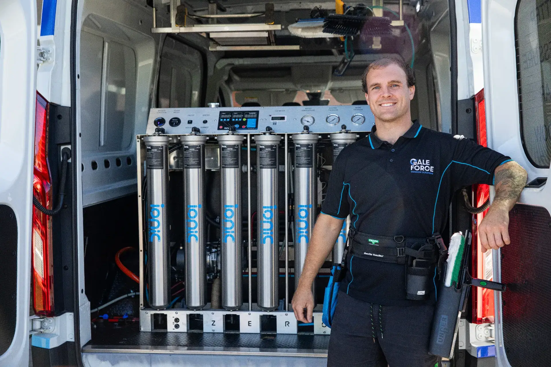 A smiling worker in a black uniform stands beside a row of metal water filtration tanks labelled "Ionic" inside the back of a white service van. Cleaning equipment hangs from his belt.