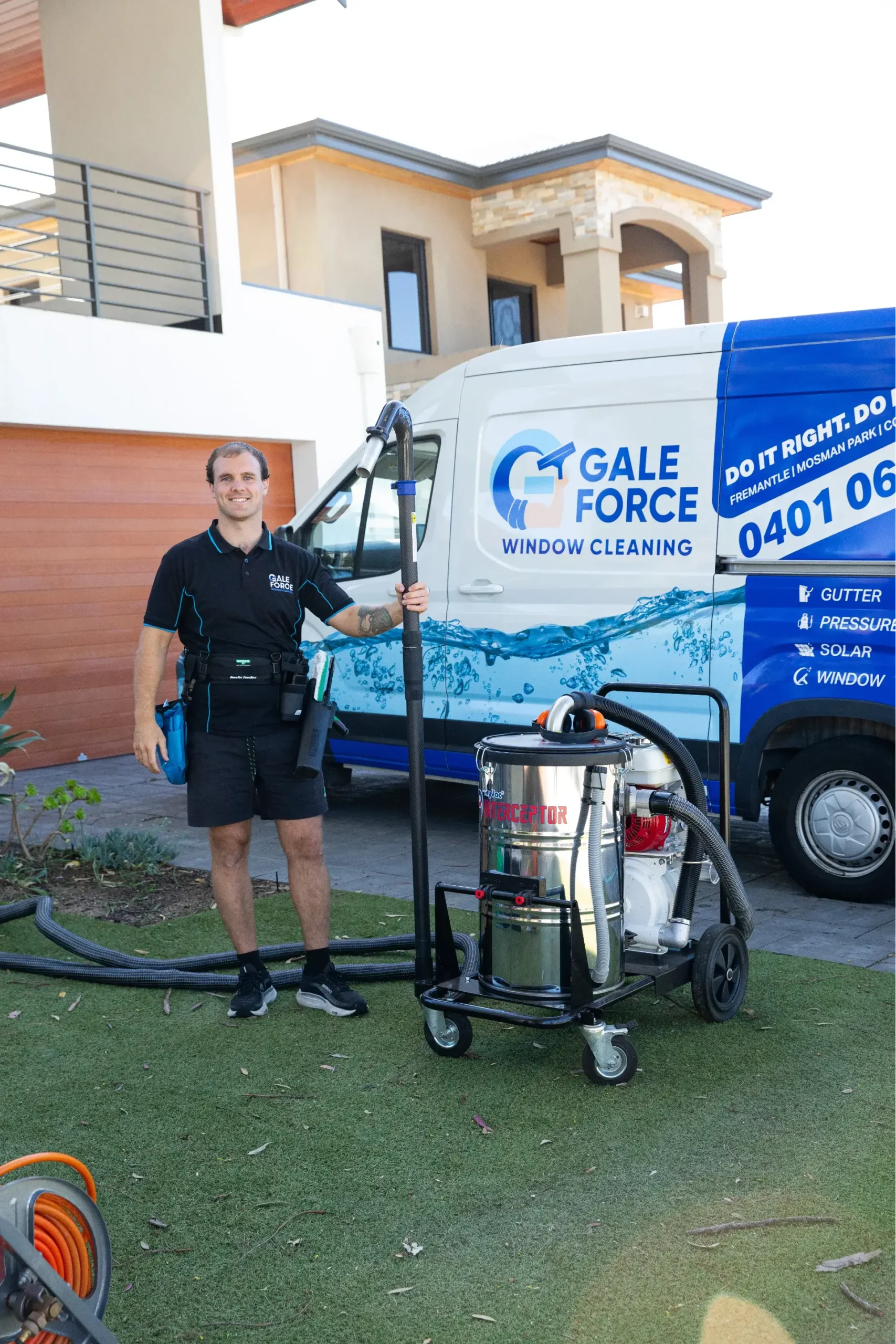 A smiling man in a black uniform stands beside a window cleaning vacuum and a blue Gale Force Window Cleaning van outside a modern house.
