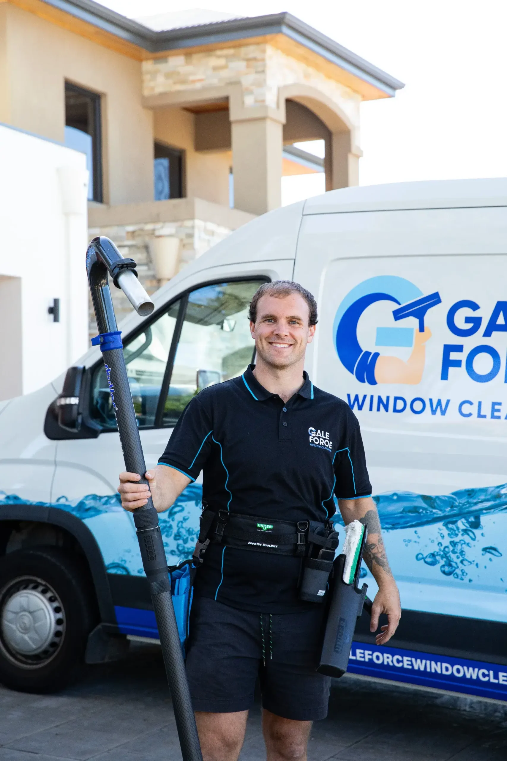 A smiling window cleaner stands in front of a van holding a long cleaning pole. He wears a black uniform and tool belt, with a modern house and branded van visible in the background.