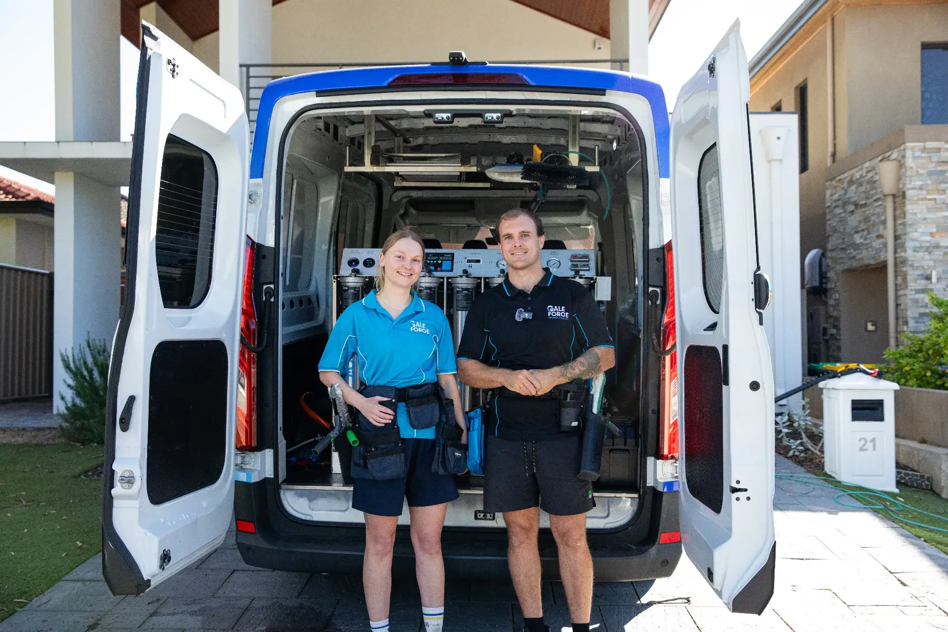 Two cleaners in uniform stand smiling in front of an open service van filled with cleaning supplies, parked outside a modern house on a sunny day.