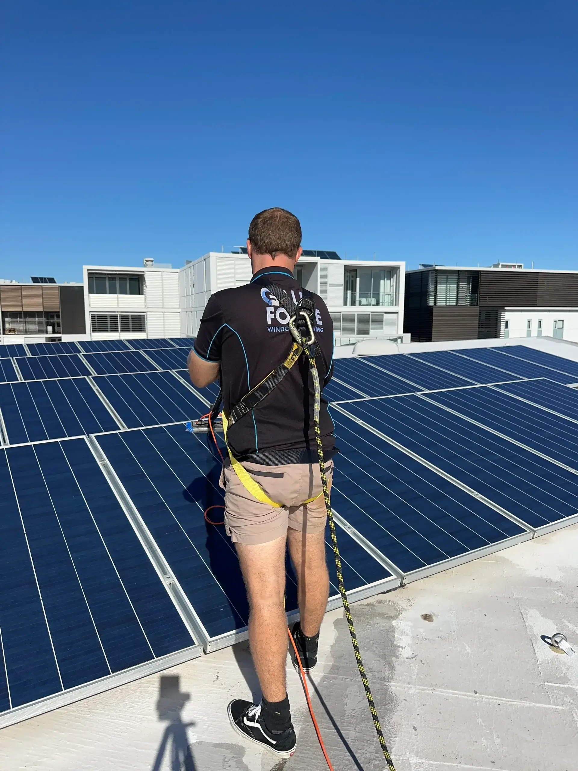 A person wearing a safety harness stands on a rooftop next to solar panels under a clear blue sky, with modern buildings visible in the background.