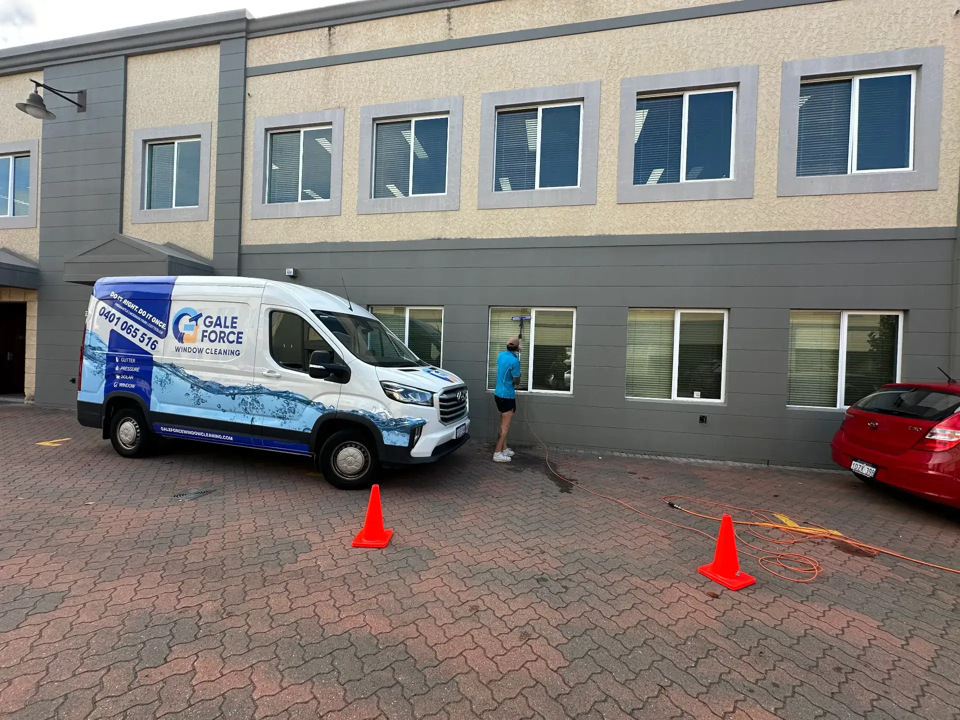 A person in blue clothing stands near a white Gale Force window cleaning van parked outside a building. Orange cones and hoses are on the ground near the van and a red car.